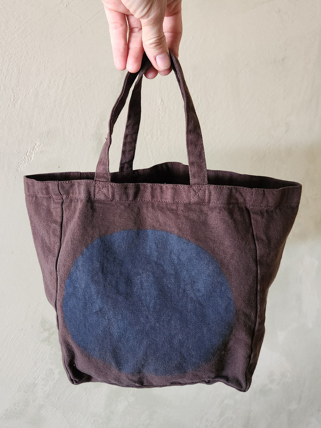 Brown tote bag with a blue circular design held by a hand against a neutral background