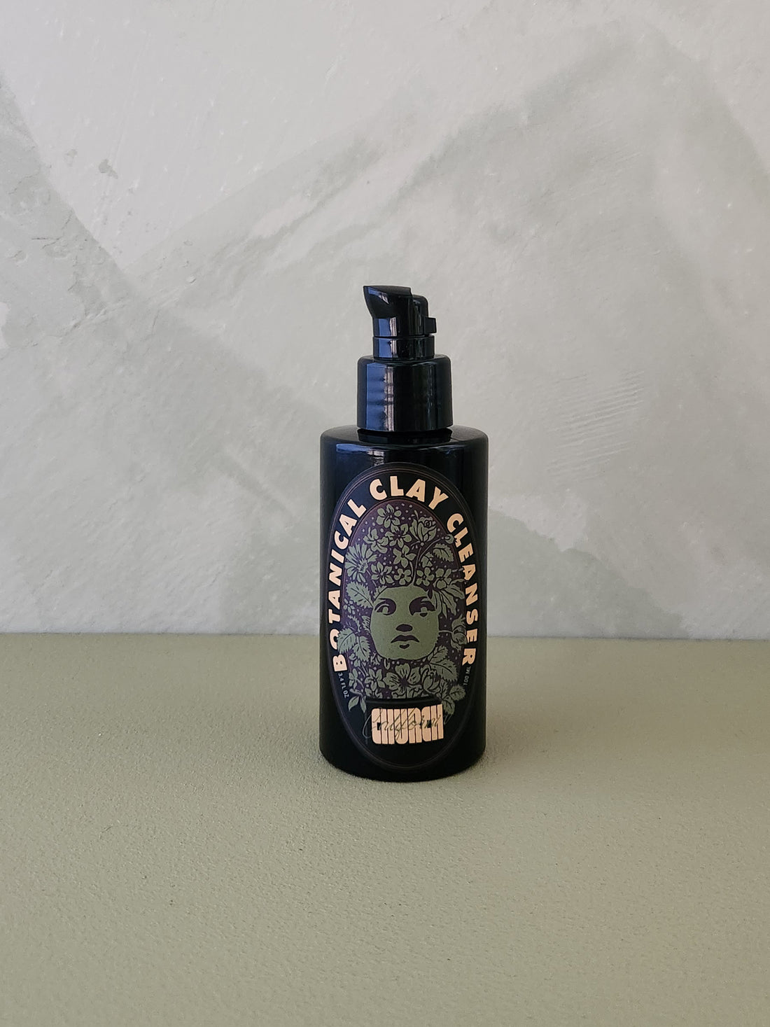 A dark pump bottle of Botanical Clay Cleanser standing on a light sage green surface against a textured light gray wall.