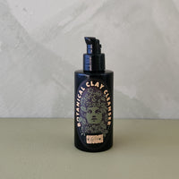 A dark pump bottle of Botanical Clay Cleanser standing on a light sage green surface against a textured light gray wall.