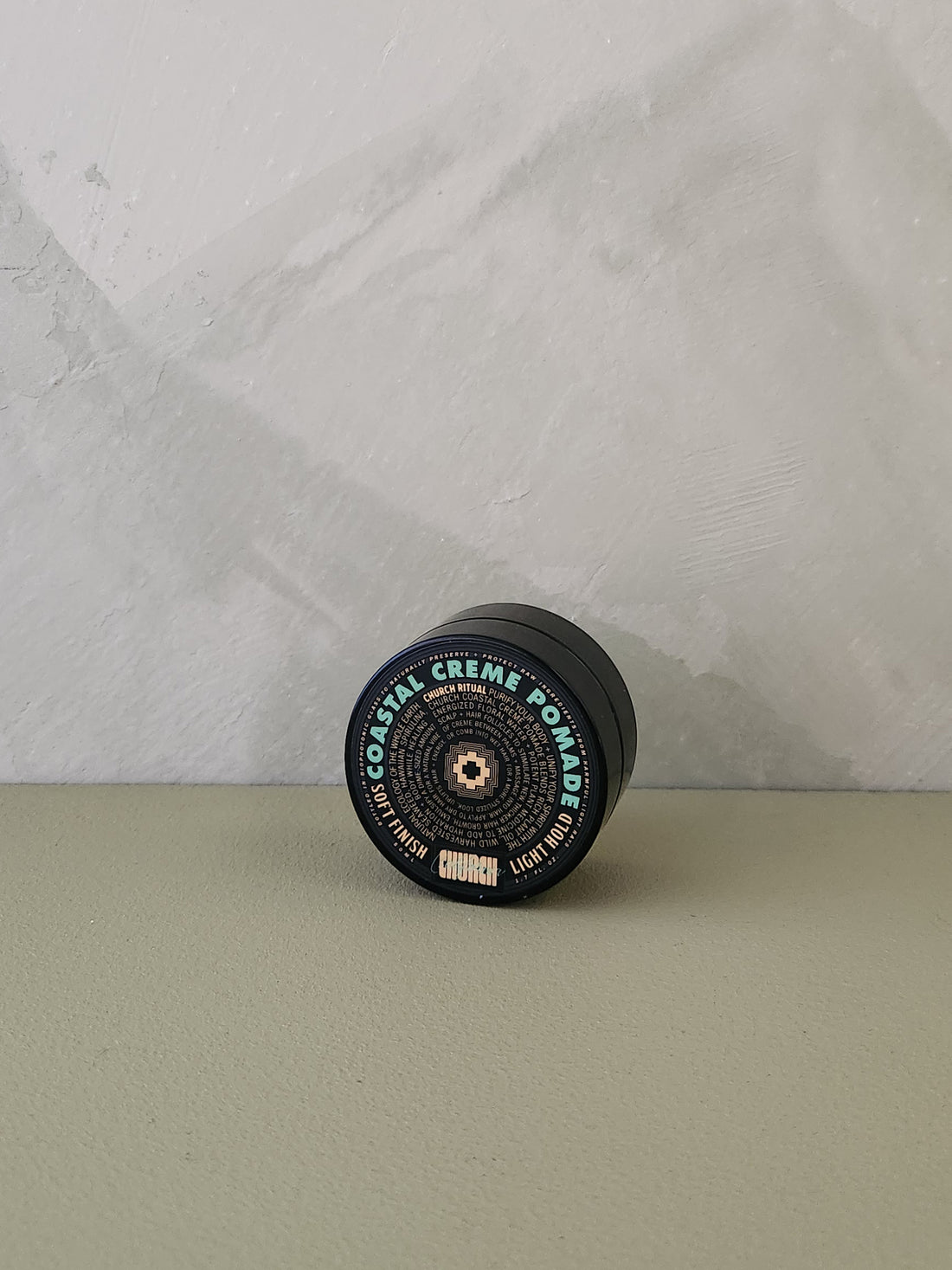 A single cylindrical cosmetic container lies on its side on a muted sage surface against a light gray textured wall. The product label reads &