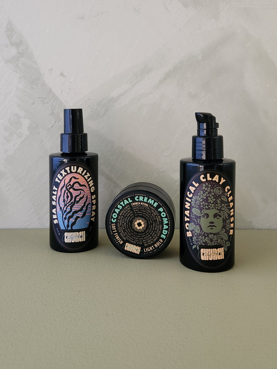 Three black haircare products from the brand Church: a texturizing spray, a clay cream, and a clay conditioner, arranged on a beige surface against a light gray marble-patterned background.