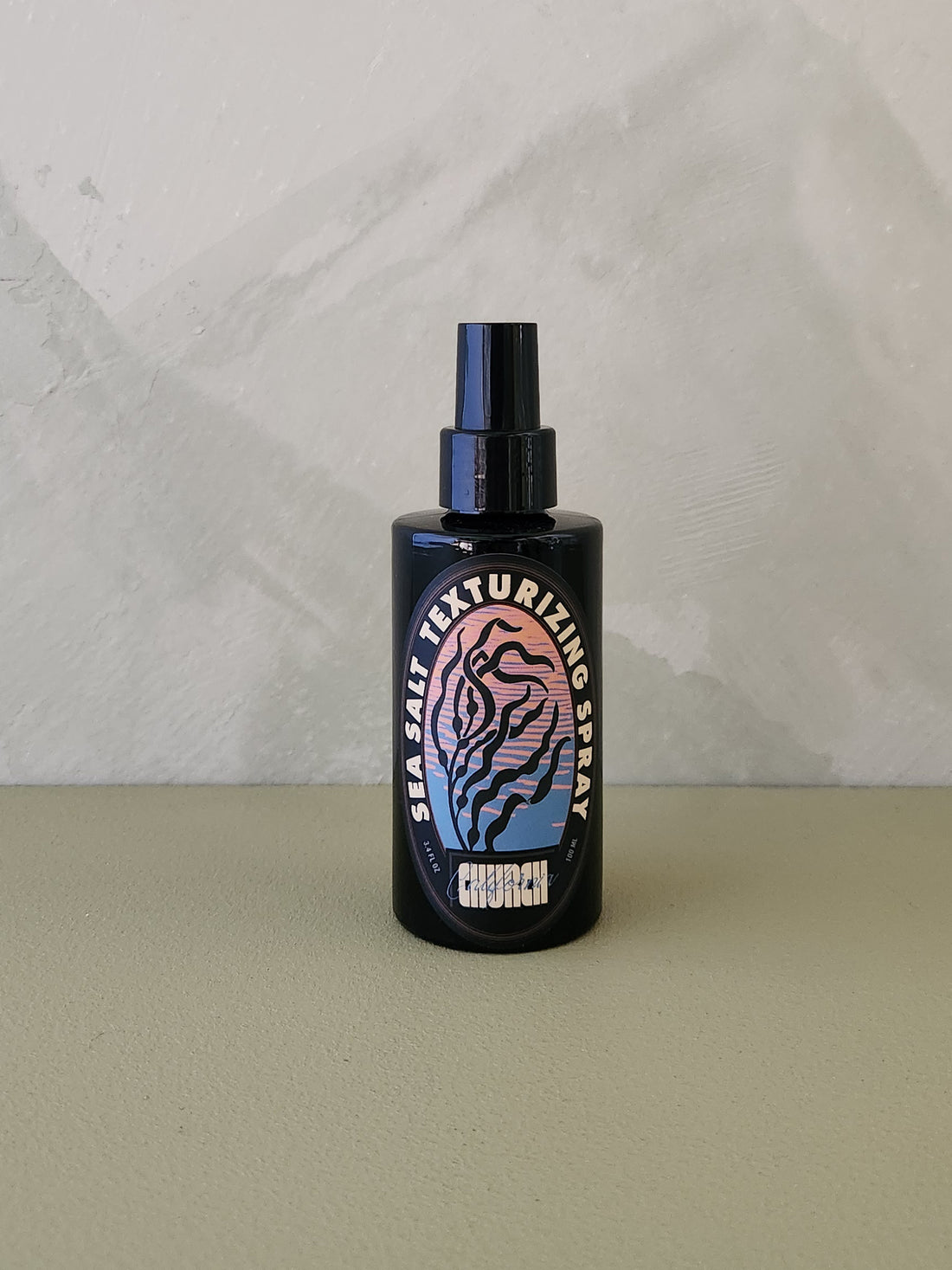 A black spray bottle of Church Sea Salt Texturizing Spray with a colorful label, set against a textured light gray wall and a beige surface.