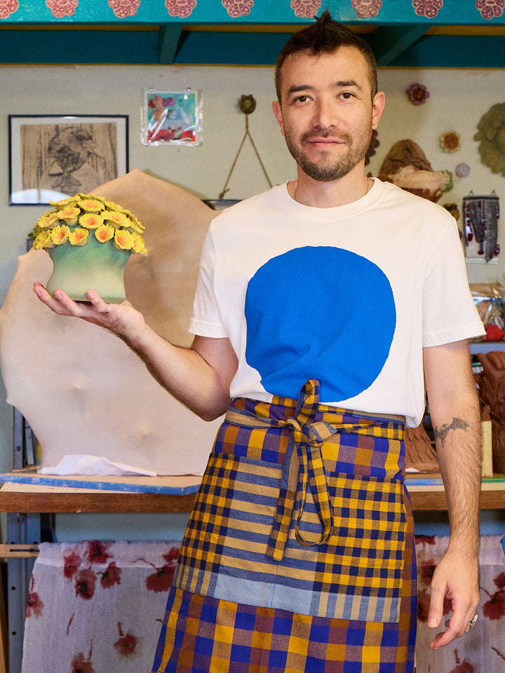Person wearing a white t-shirt with a blue circle design and a plaid apron, standing in a workshop.
