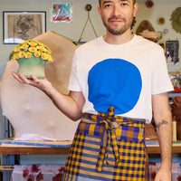 Person wearing a white t-shirt with a blue circle design and a plaid apron, standing in a workshop.