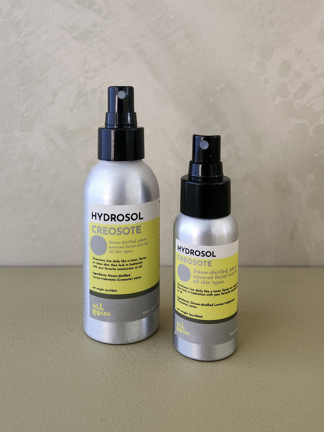 Two silver Hydrosol Creosote bottles with black spray tops and yellow labels, standing on a beige surface against a textured beige wall.