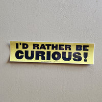 A yellow bumper sticker with bold black text reading 'I'D RATHER BE CURIOUS!' mounted on a textured light gray wall.