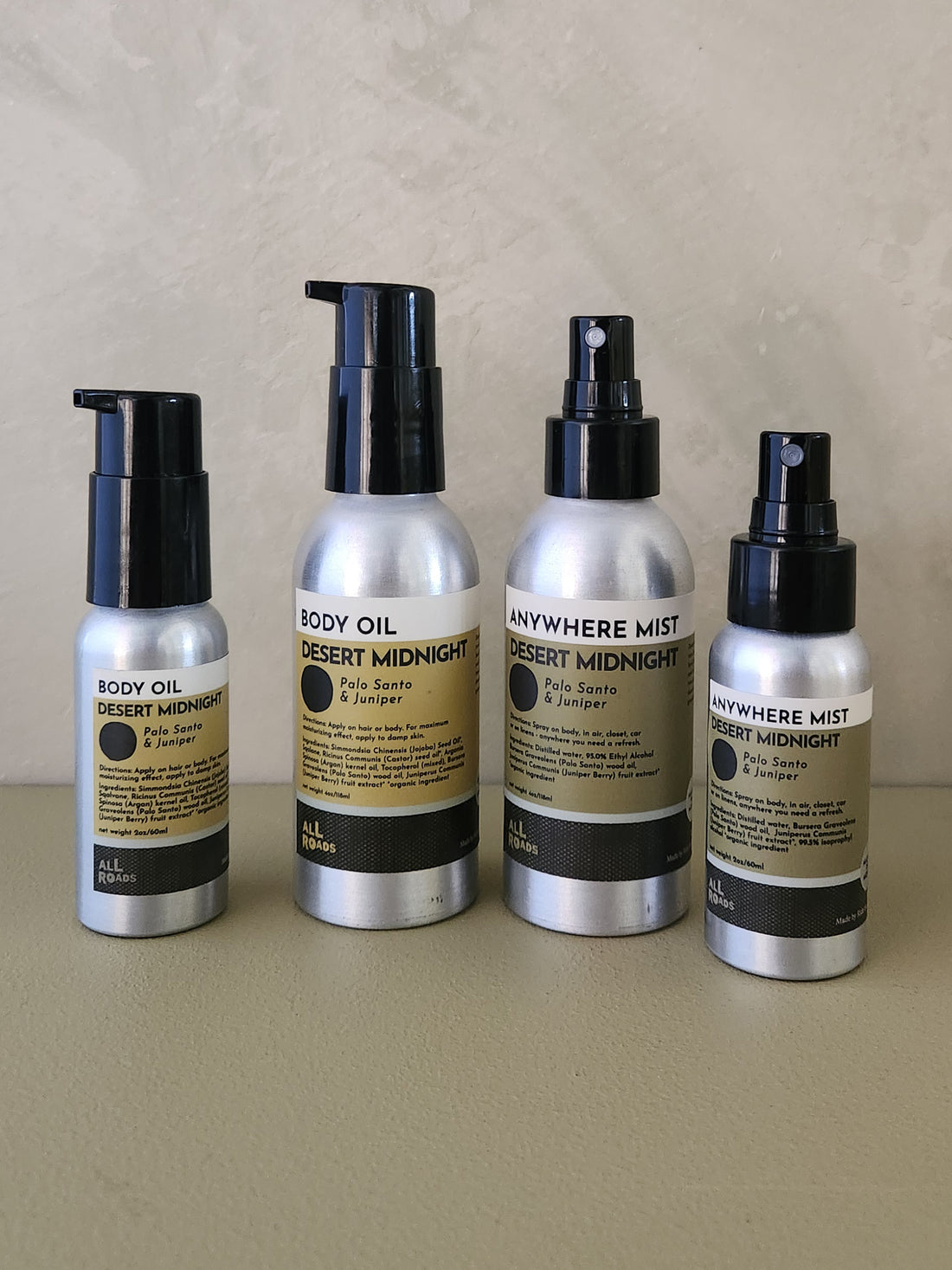 Four fragrance products from All Roads and Body Care lined up on a beige surface against a textured beige wall. The set includes Desert Midnight Body Oil, Desert Midnight Body Oil, Desert Midnight Body Mist, and Desert Midnight Anywhere Mist in silver bottles with black caps and tan labels.