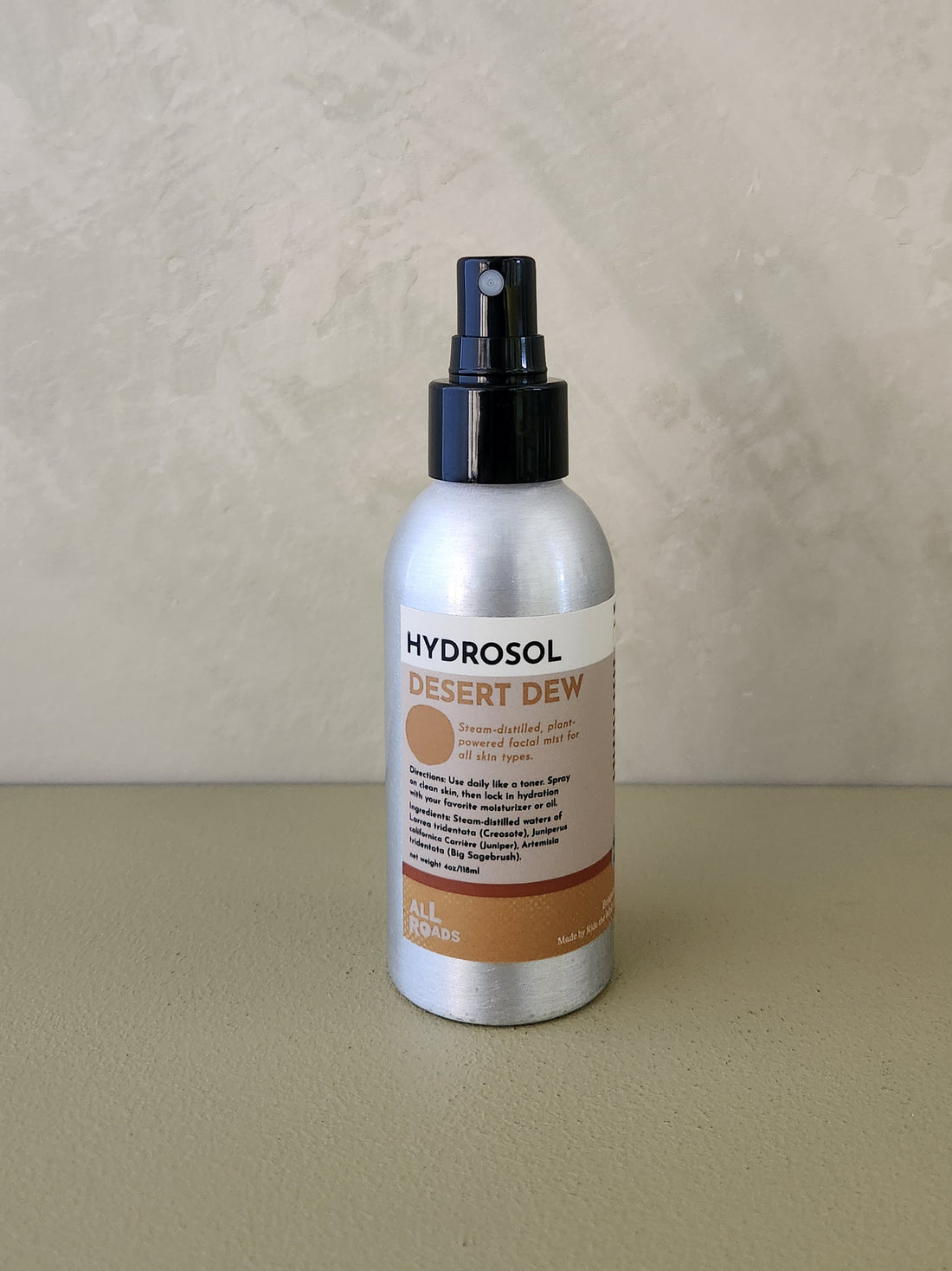 A bottle of Hydrosol Desert Dew room spray with a silver metallic body, black spray nozzle, and a beige and brown label, set against a textured beige background.