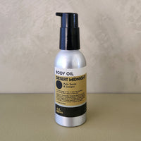 A bottle of body oil with a black pump and a silver bottle body, featuring a beige and black label that reads 'DESERT MIDNIGHT' and 'Palm Oils / Juglans', set against a textured beige background.