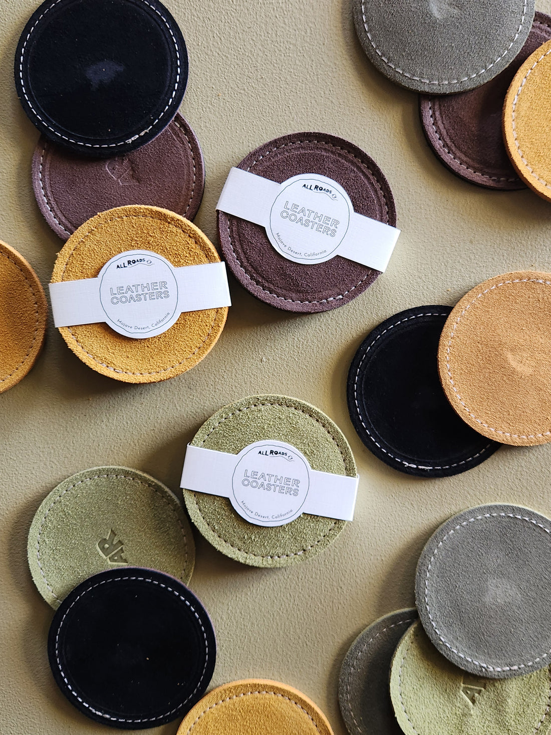 A flat-lay arrangement of circular leather coasters in multiple colors, including black, brown, mustard yellow, sage green, olive, tan, and gray, each with white stitched edges and a white paper band label reading &