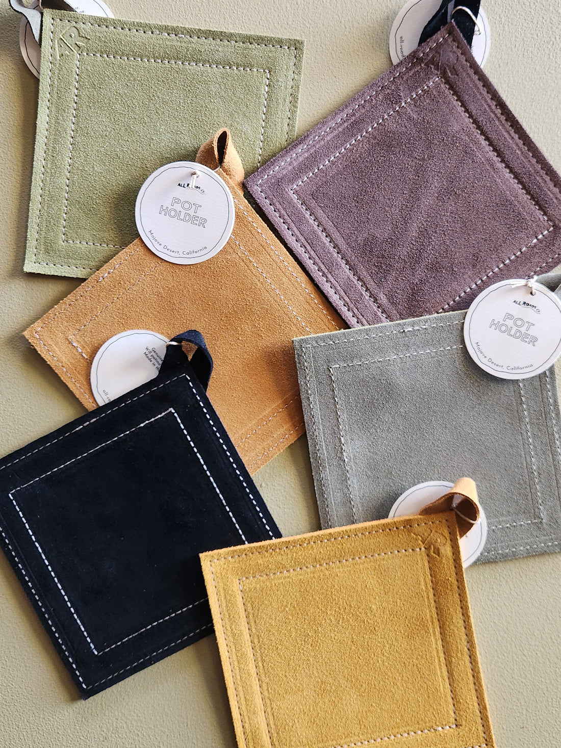 Six square suede-like pouches or coasters in assorted muted colors, arranged in a flat lay on a beige surface. Each item has a small loop tab, a round white hangtag reading &