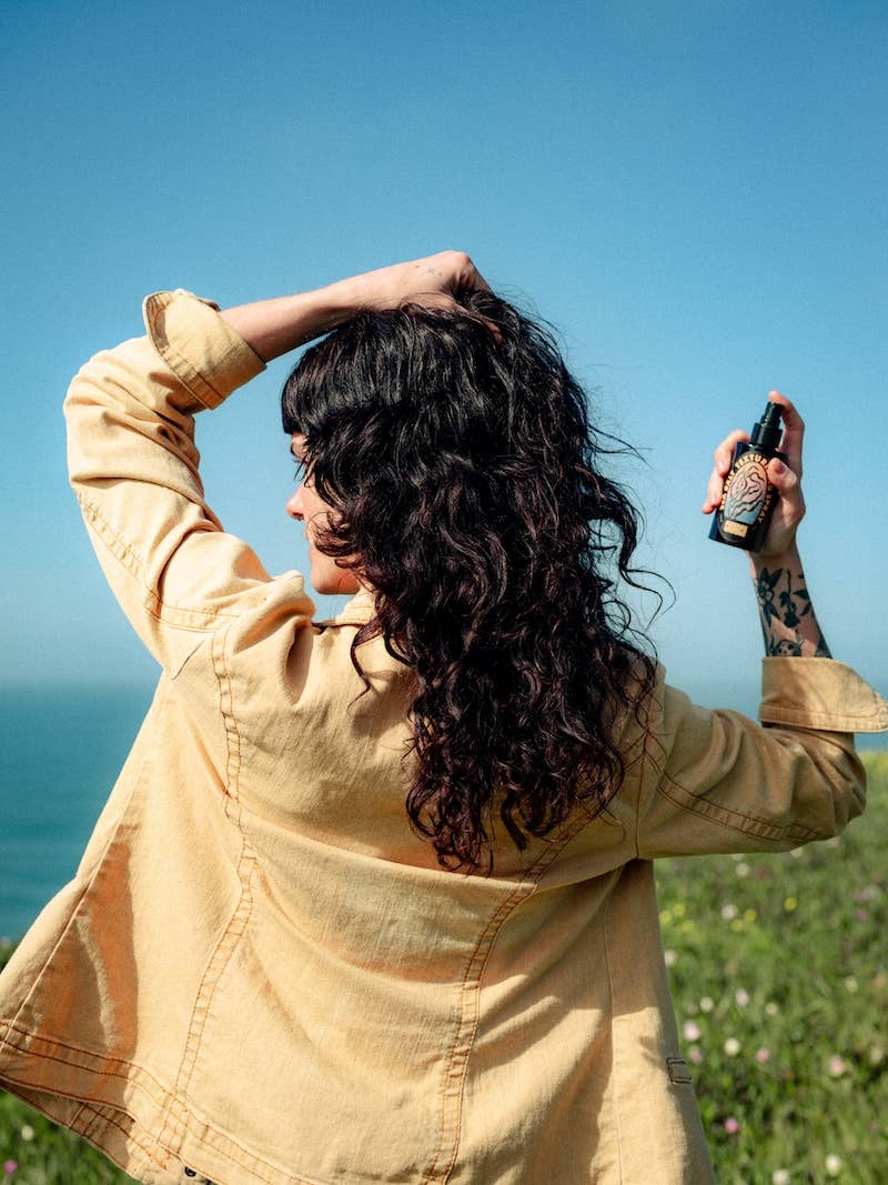 A woman with long, curly black hair is shown from behind outdoors, holding a small black spray bottle to her hair. She is wearing a light beige jacket, with a blue sky and green grass in the background.