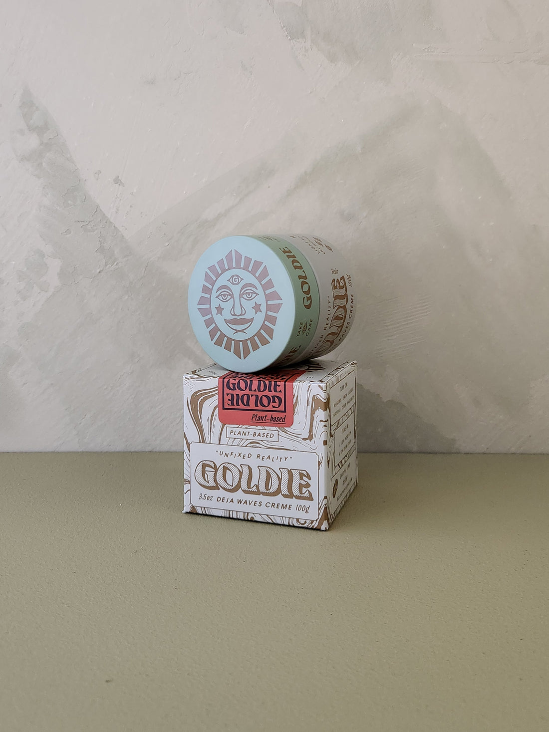 A jar of Goldie Goldie hair pomade resting on top of its branded cardboard box, set against a textured light gray wall and a muted olive-green surface.