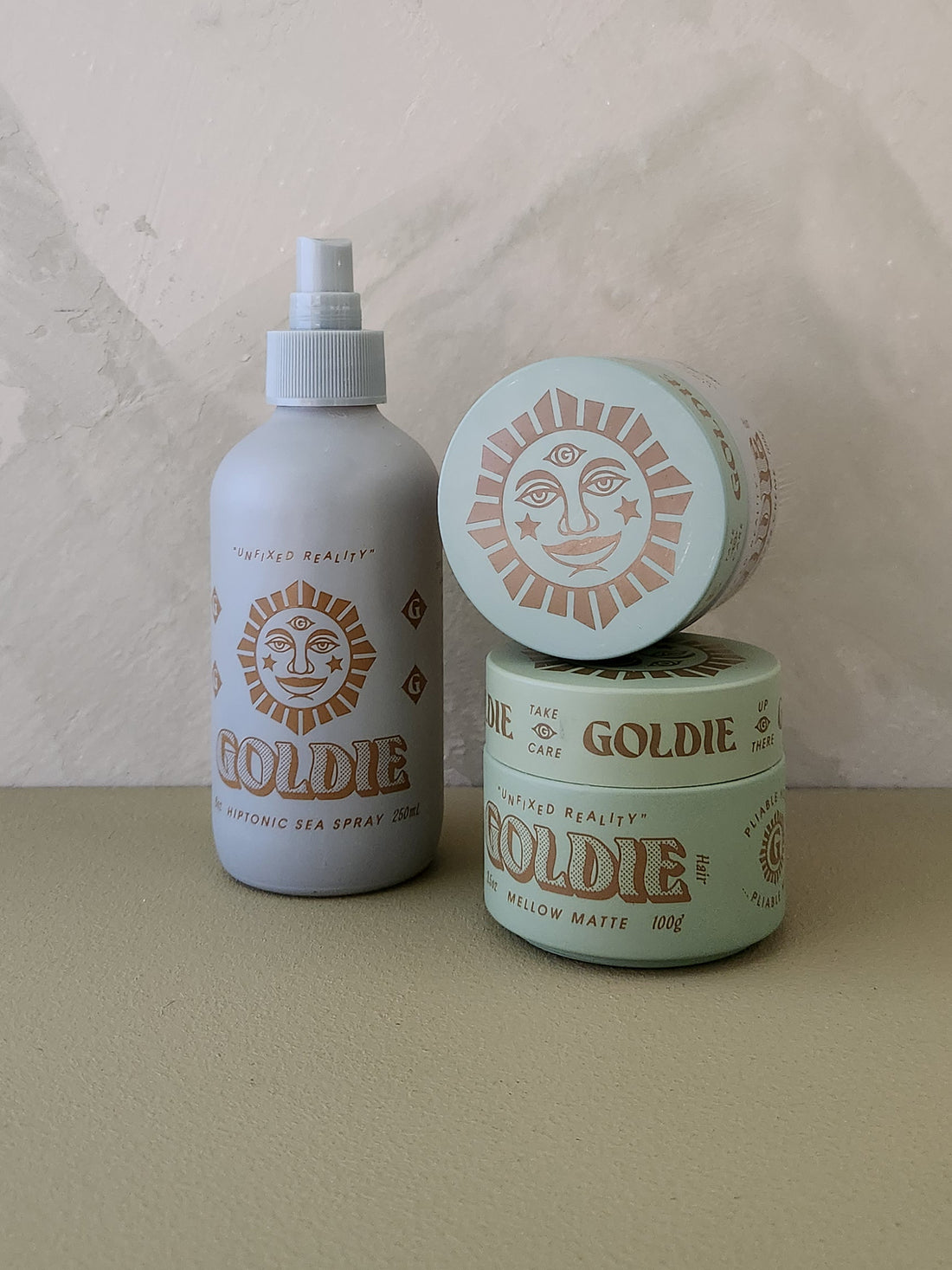 Three Goldie brand hair care containers on a beige surface against a textured beige wall: a matte spray bottle, a round jar, and a smaller round tin stacked on top. The bottle is light gray with tan sun graphics, while the jars are pale mint green with tan sun graphics.