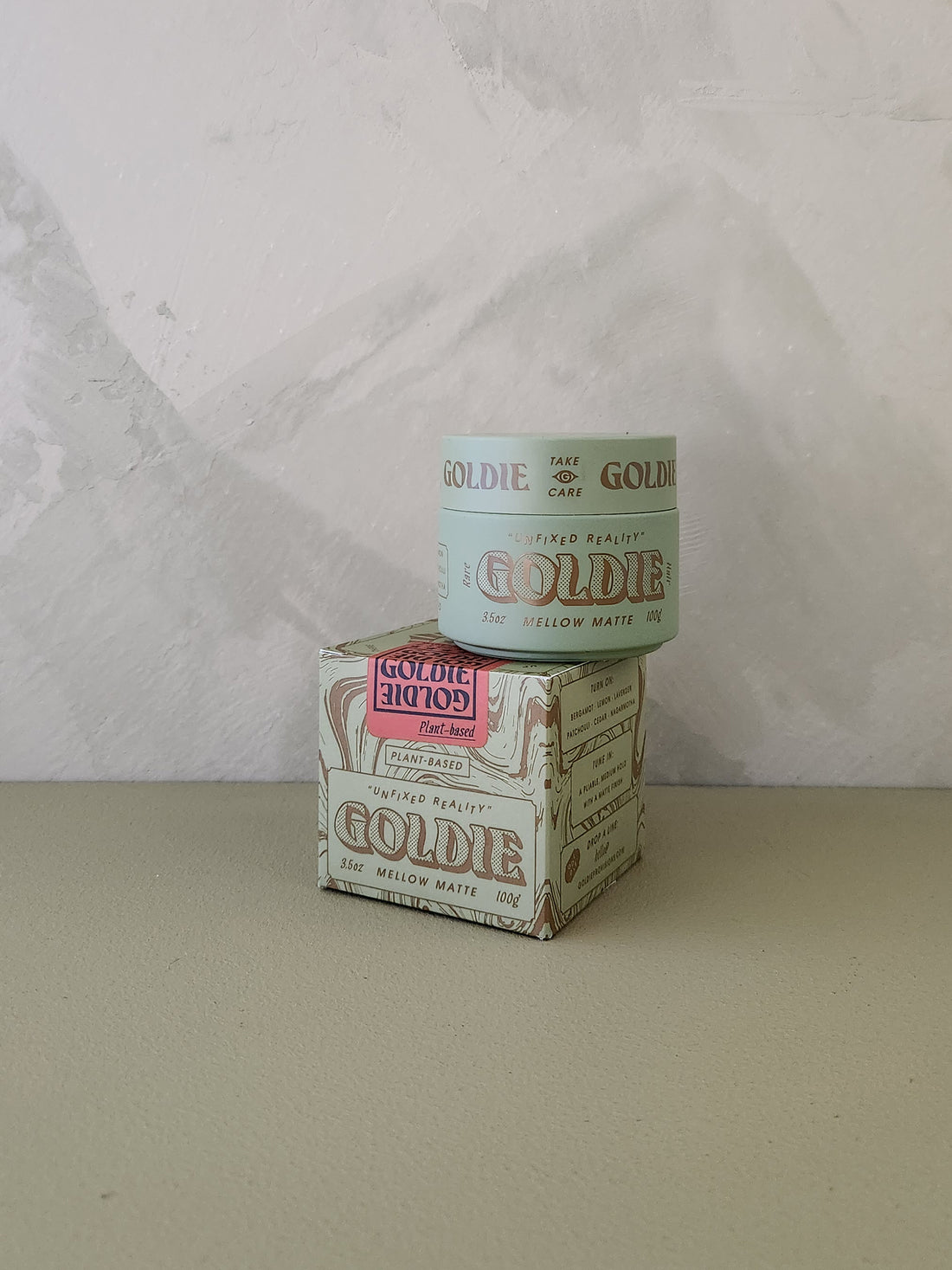 A small jar of Goldie Mellow Matte hair pomade sits on top of its matching printed box, photographed against a light textured wall and neutral tabletop.