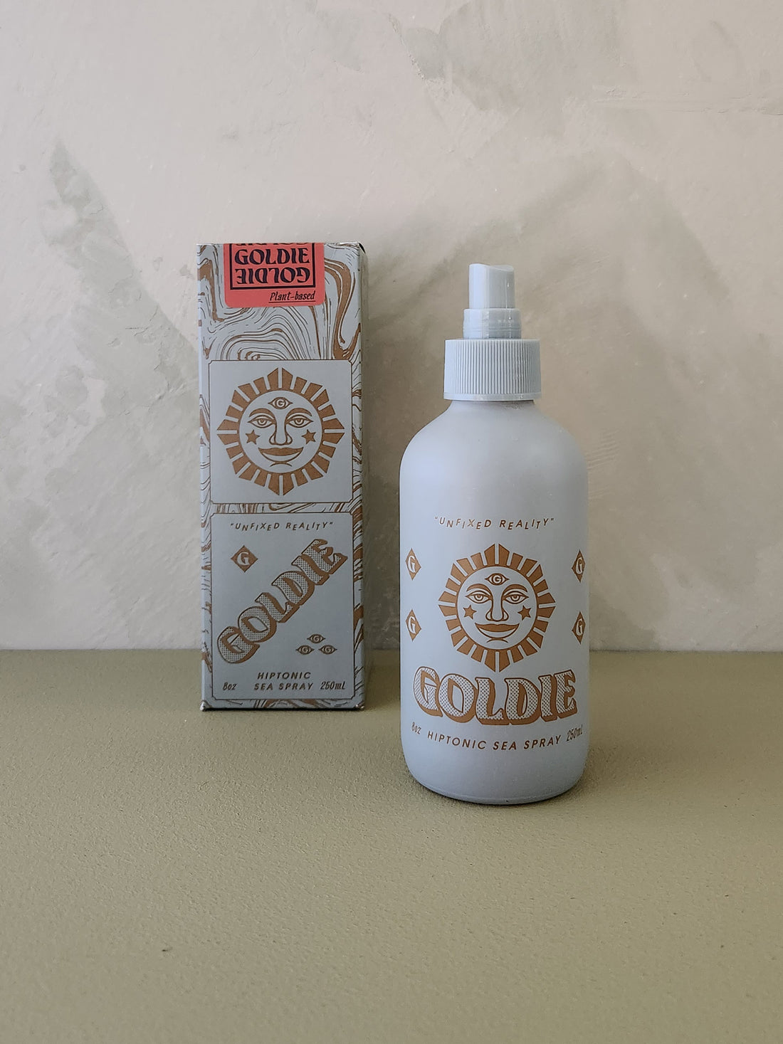 A bottle of Goldie Sea Spray next to its retail box, both featuring a gold sun face logo on a white background, set against a textured beige wall and surface.