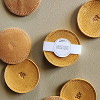 A set of round tan leather coasters with white stitching and embossed 'AR' initials, arranged on a textured beige surface. One coaster in the center is wrapped with a white paper band labeled 'ALL ROADS LEATHER COASTERS'.