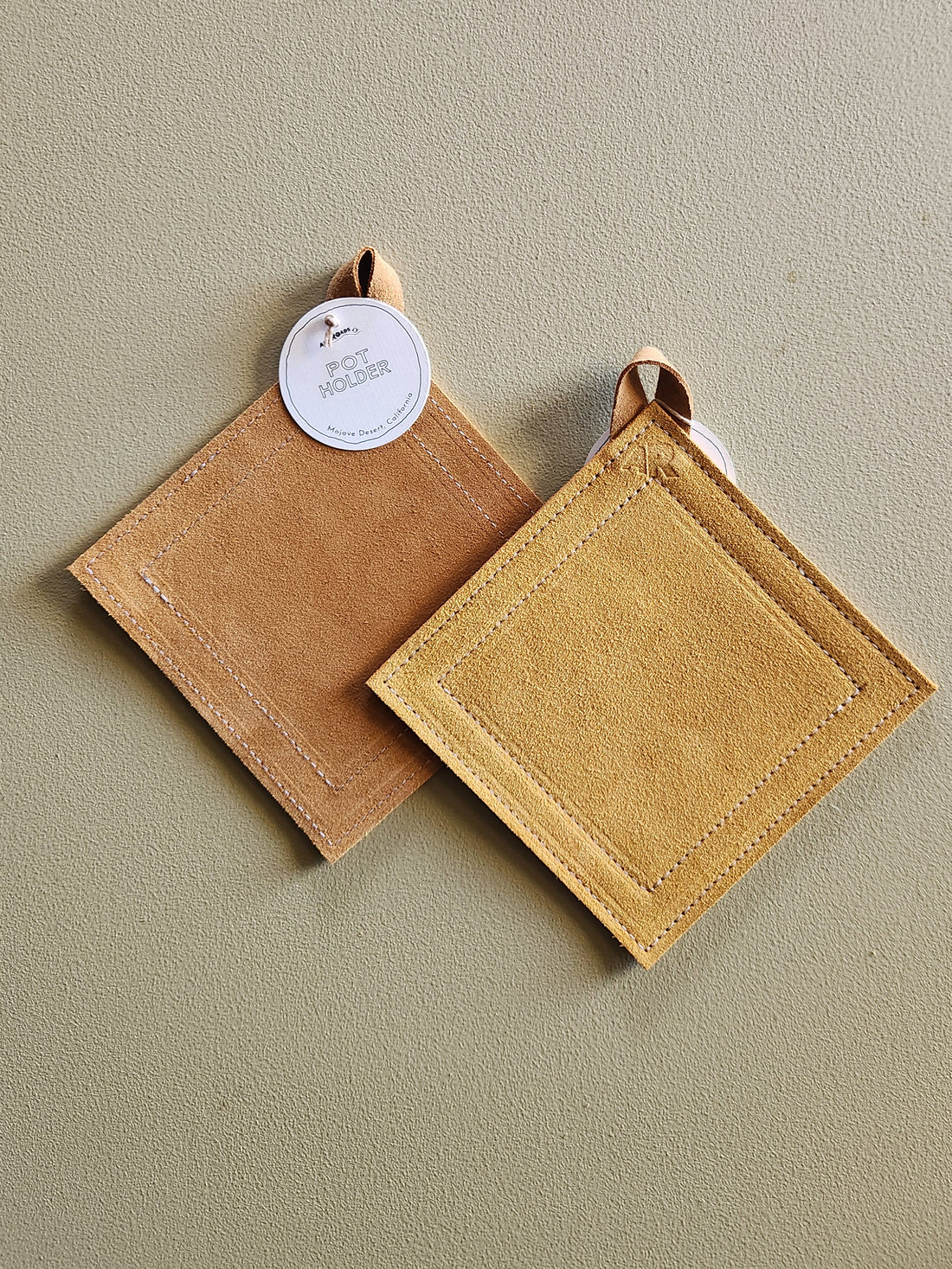 Two square suede-like potholders in tan and mustard yellow, shown overlapping on a neutral beige background. One has a white circular hang tag.