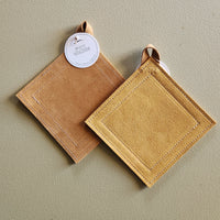 Two square suede-like potholders in tan and mustard yellow, shown overlapping on a neutral beige background. One has a white circular hang tag.