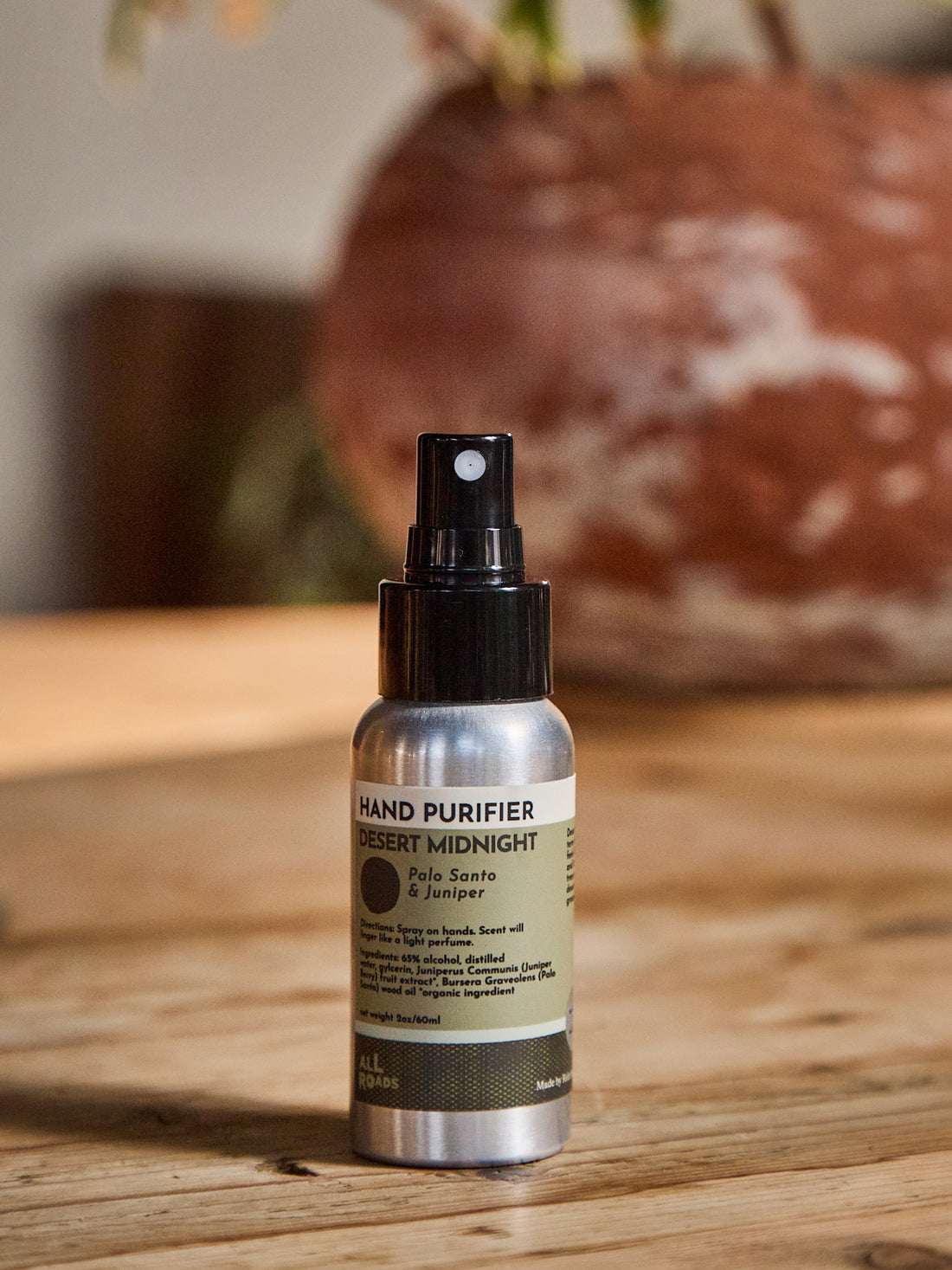 Hand purifier in a  spray bottle with a black spray top and a silver and olive green label, placed on a wooden surface with a blurred rustic background.