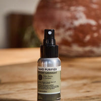 Hand purifier in a  spray bottle with a black spray top and a silver and olive green label, placed on a wooden surface with a blurred rustic background.