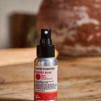 A small hand sanitizer bottle labeled 'HAND PURIFIER DESERT ROSE' with a black spray top and silver bottle, standing on a wooden surface with a blurred rustic background.