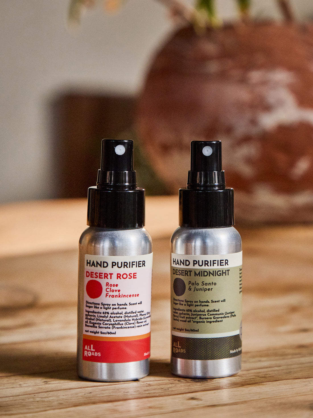 Two hand purifier spray bottles standing on a wooden surface, one labeled Desert Rose with a red label and one labeled Desert Midnight with a dark olive label. A blurred ceramic pot and indoor setting are in the background.