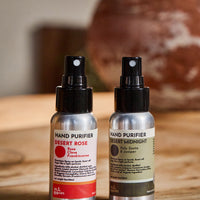 Two hand purifier spray bottles standing on a wooden surface, one labeled Desert Rose with a red label and one labeled Desert Midnight with a dark olive label. A blurred ceramic pot and indoor setting are in the background.