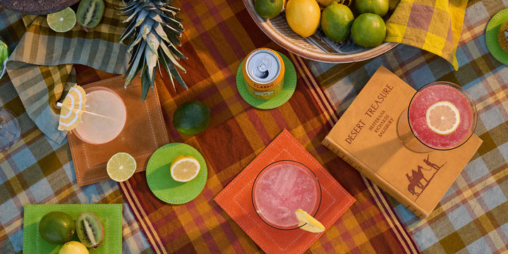 Cocktail setup with lime slices, a candle, and a book on a plaid tablecloth.