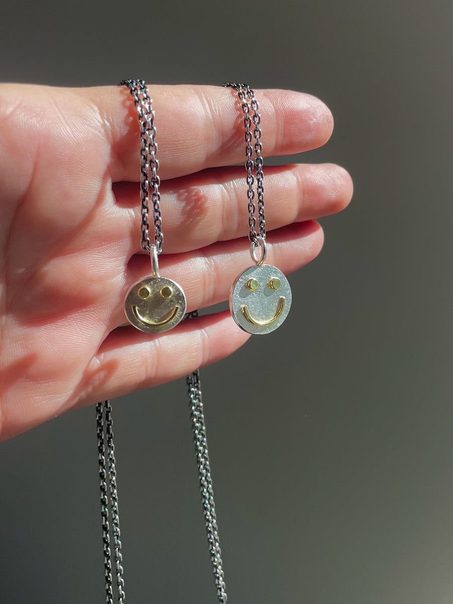 Two smiley face pendants on chains held in a hand against a neutral background