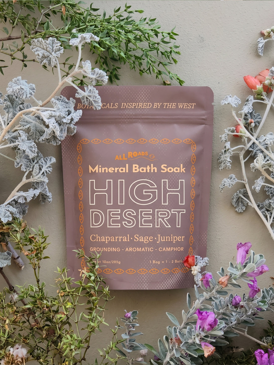 A stand-up pouch of All Roads Mineral Bath Soak, High Desert variant, styled with botanical sprigs and flowers against a neutral beige background.