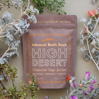 A stand-up pouch of All Roads Mineral Bath Soak, High Desert variant, styled with botanical sprigs and flowers against a neutral beige background.
