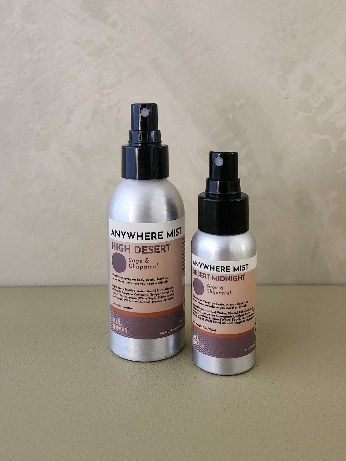 Two silver spray bottles of Anywhere Mist body mist, one labeled High Desert and one labeled Desert Midnight, standing on a beige surface against a textured beige wall.