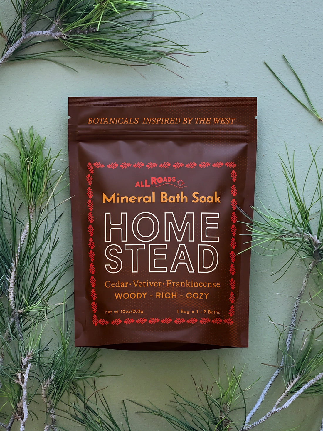 A stand-up pouch of All Roads Home Stead Mineral Bath Soak is centered against a sage green background, surrounded by pine branches. The pouch is dark brown with cream and red text and border details.