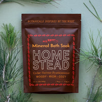 A stand-up pouch of All Roads Home Stead Mineral Bath Soak is centered against a sage green background, surrounded by pine branches. The pouch is dark brown with cream and red text and border details.