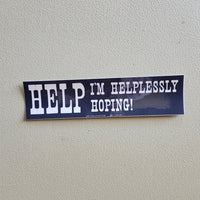 A dark blue bumper sticker with white text that reads 'HELP I'M HELPLESSLY HOPING!' on a textured beige w