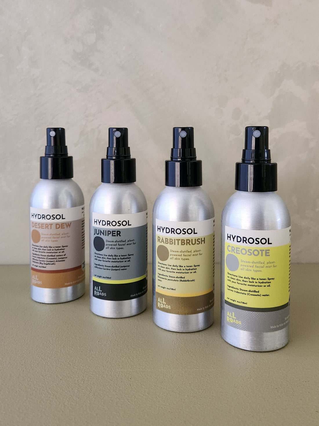 Four Hydrosol room spray bottles lined up on a beige surface against a textured beige wall. The bottles are silver aluminum with black spray tops and feature labels in warm neutral tones: Desert Dew (tan), Juniper (gray/black), Rabbitbrush (gold/brown), and Creosote (yellow/gray).
