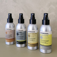 Four Hydrosol room spray bottles lined up on a beige surface against a textured beige wall. The bottles are silver aluminum with black spray tops and feature labels in warm neutral tones: Desert Dew (tan), Juniper (gray/black), Rabbitbrush (gold/brown), and Creosote (yellow/gray).