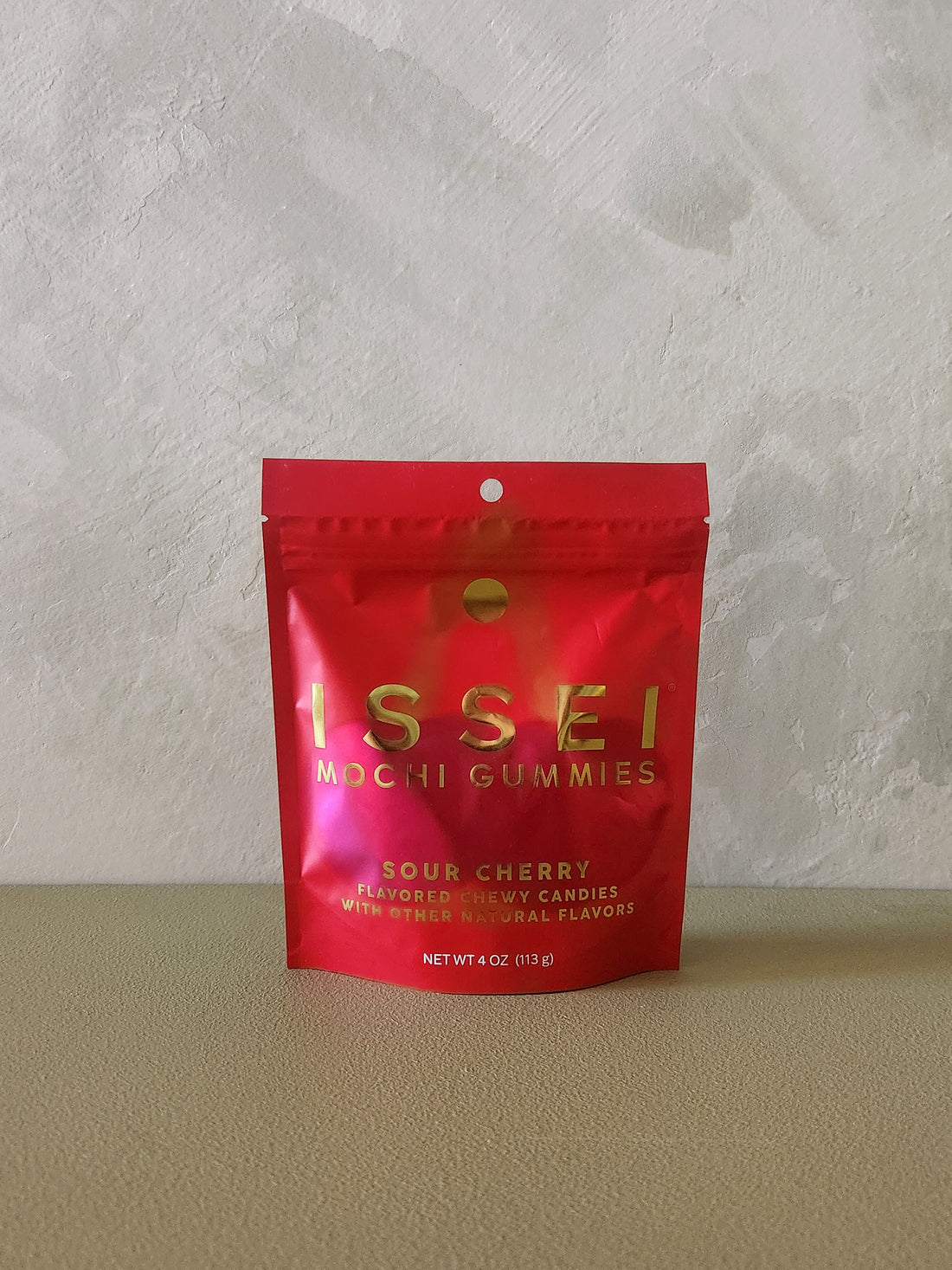 A red stand-up pouch of Issei Mochi Gummies, Sour Cherry flavor, standing on a beige surface against a textured light gray wall.