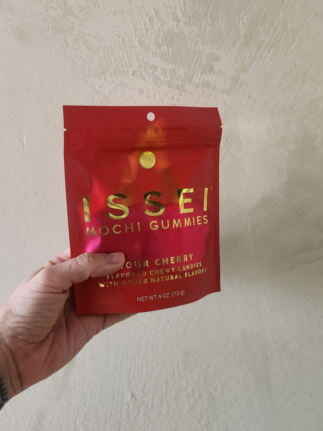 A hand holding a red stand-up pouch of Issei Mochi Gummies, Sour Cherry flavor, against a textured beige wall.