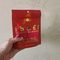 A hand holding a red stand-up pouch of Issei Mochi Gummies, Sour Cherry flavor, against a textured beige wall.