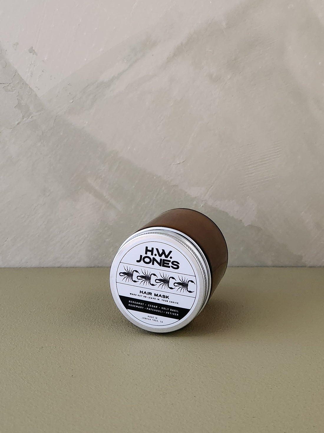 A glass jar of H.W. Jones Hair Mask lies on its side on a textured beige surface against a neutral gray wall.
