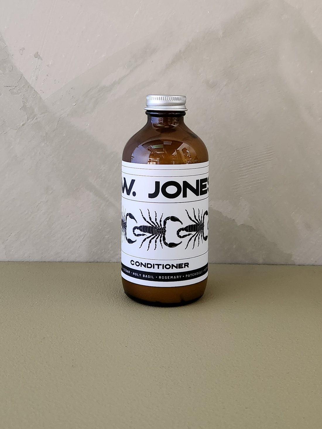 An amber glass bottle of Jones Conditioner with a white label and silver cap, set against a textured beige wall and surface.