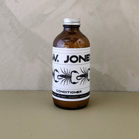 An amber glass bottle of Jones Conditioner with a white label and silver cap, set against a textured beige wall and surface.