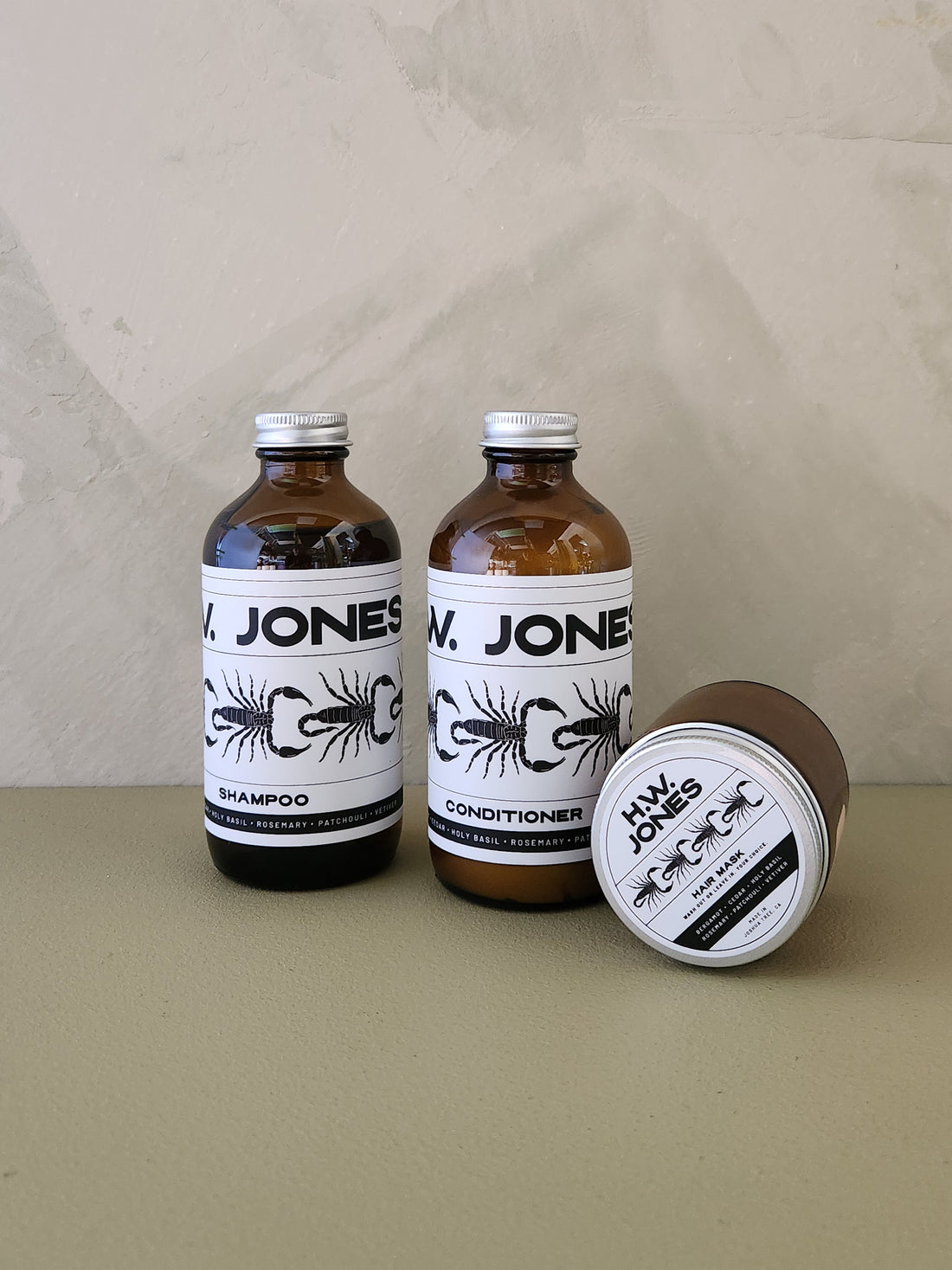 Two amber glass bottles of Jones shampoo and conditioner with white labels, alongside a matching brown jar lid, set against a neutral beige background.