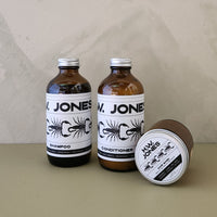 Two amber glass bottles of Jones shampoo and conditioner with white labels, alongside a matching brown jar lid, set against a neutral beige background.