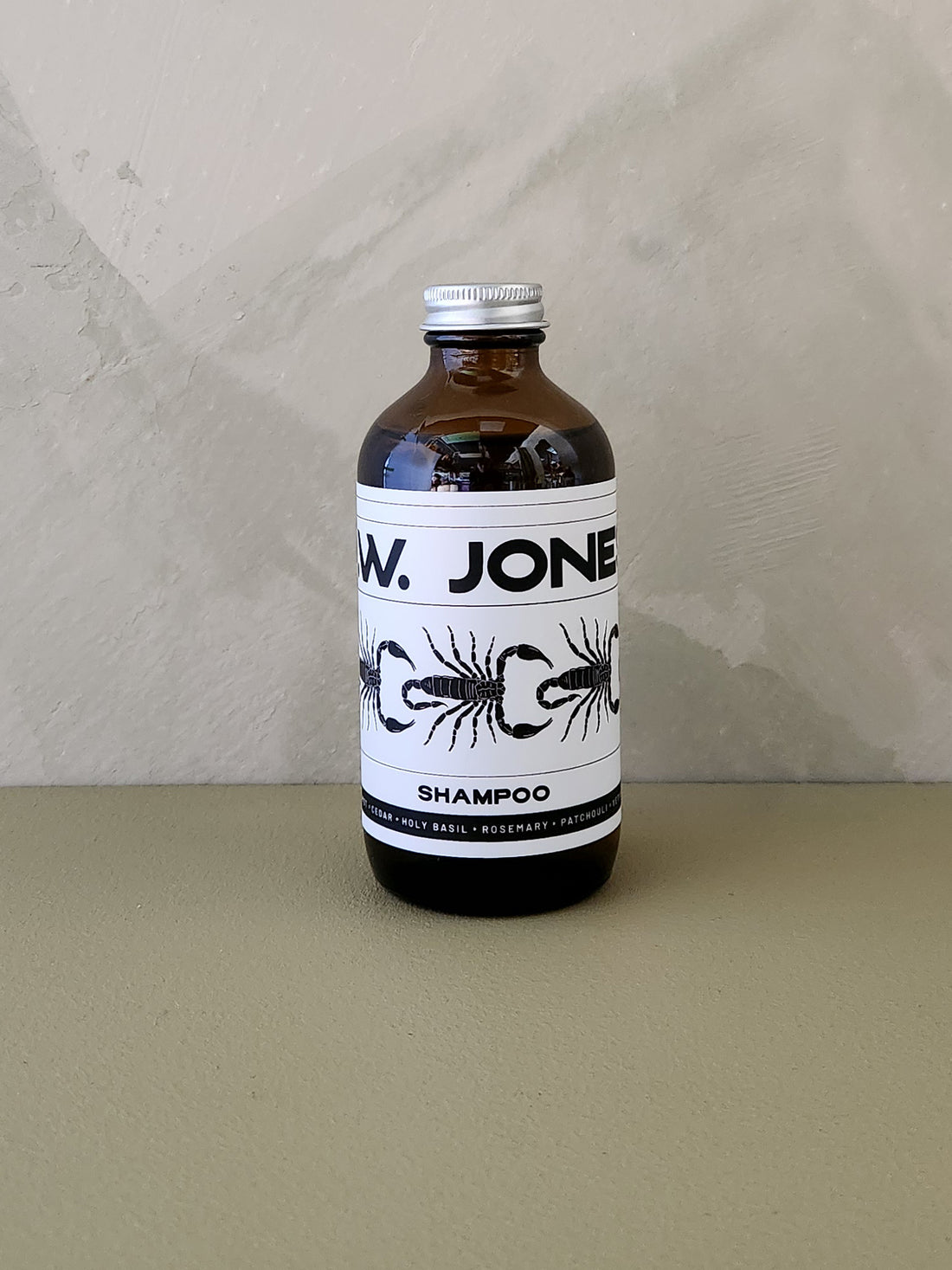 An amber glass bottle of W. Jones Shampoo with a white label and silver cap, set against a neutral textured beige background.