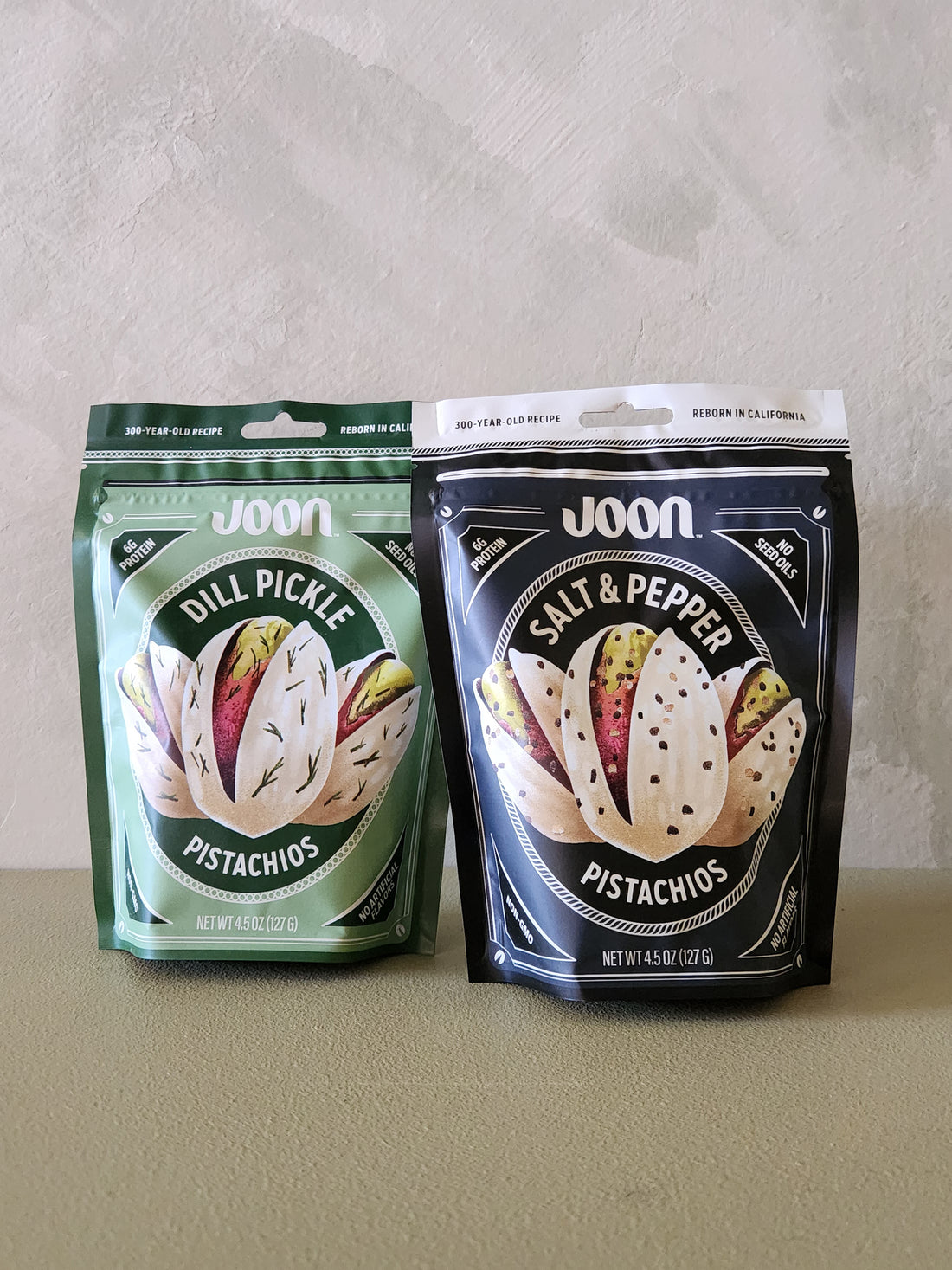 Two packages of Joon pistachios, one green and one black, on a neutral background.
