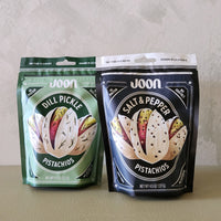 Two packages of Joon pistachios, one green and one black, on a neutral background.
