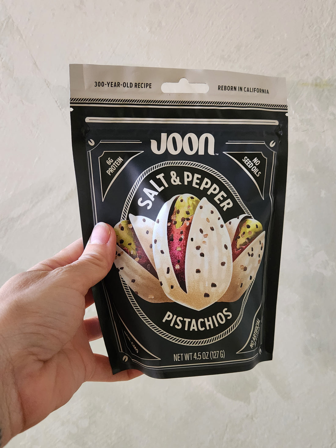 Hand holding a package of Joon Salt & Pepper Pistachios against a neutral background